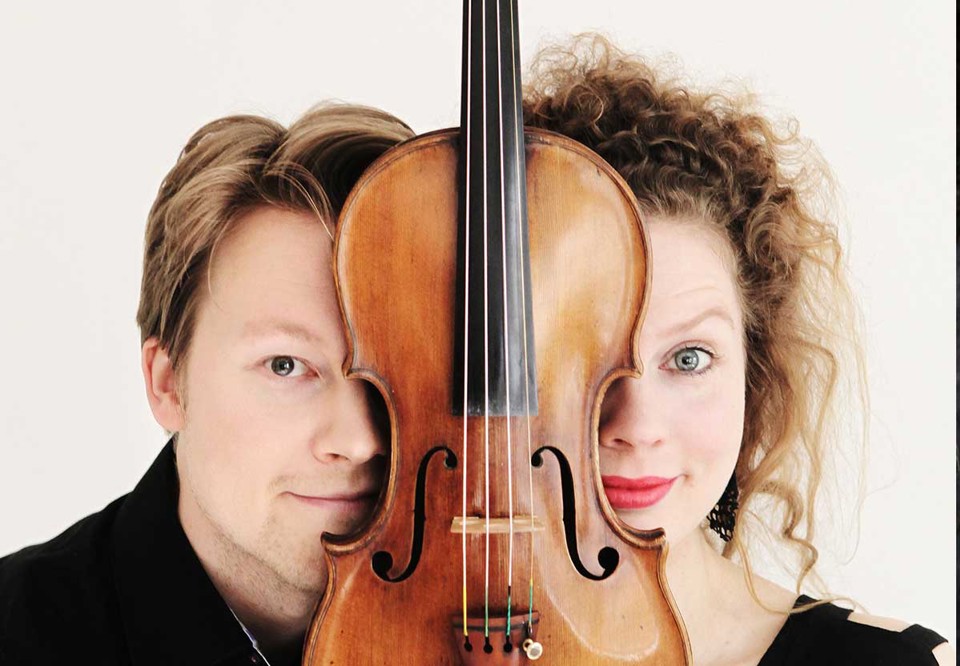 En man och en kvinna tittar in i kameran, mellan sig har de ett instrument. 