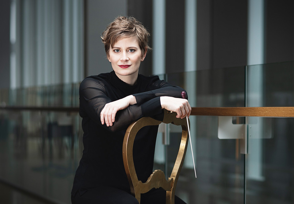 Kvinna som sitter på en stol med en dirigentpinne i handen. Fotografi.